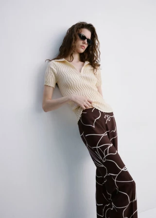 Adult female fashion model with brown curly hair poses in polo shirt and patterned trousers against a white wall in soft daylight.