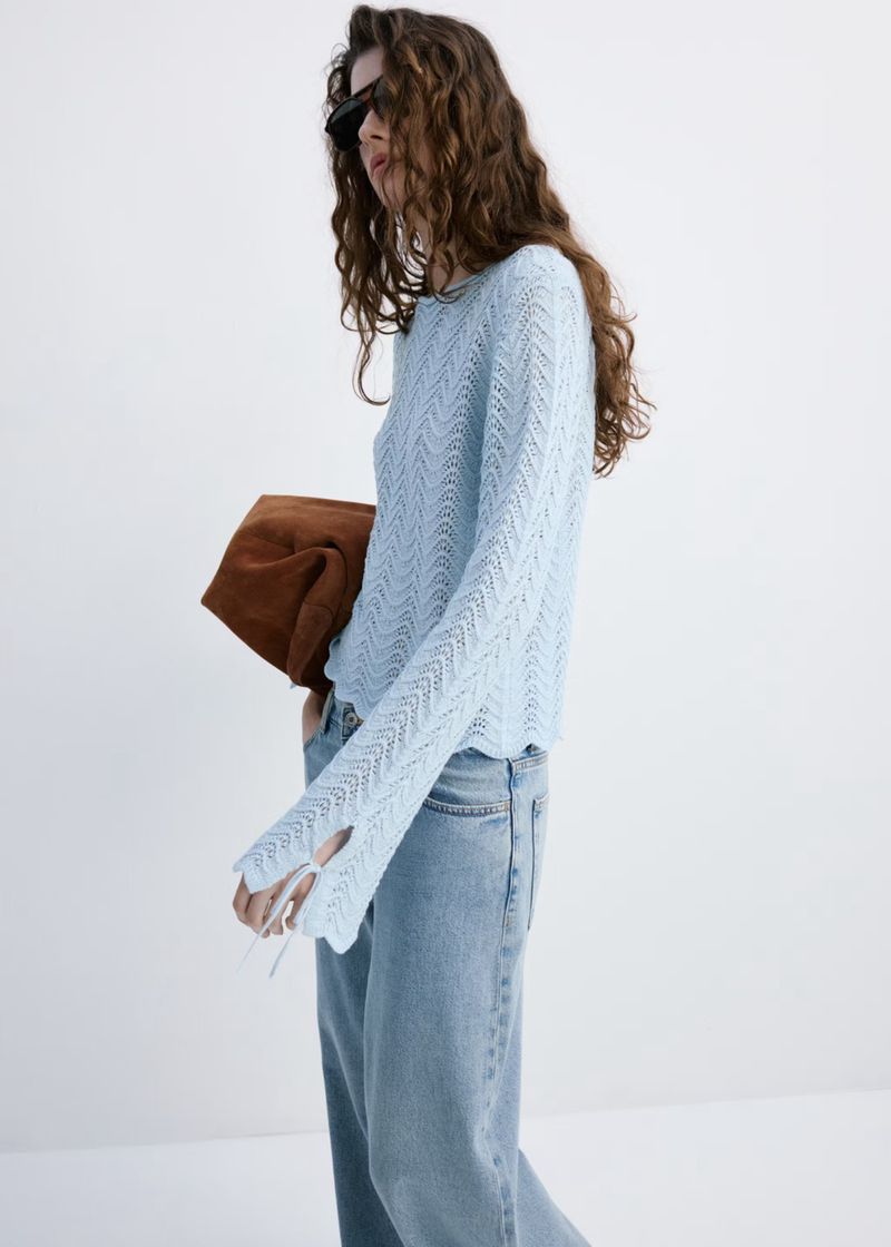 A woman in a light blue knit sweater and jeans poses against a white background, carrying a brown bag over her shoulder.