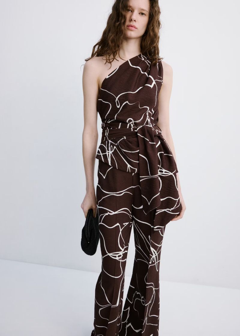 A slender woman poses in a brown one-shoulder jumpsuit with a white line pattern, holding a black clutch against a plain white background.
