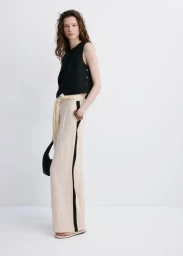 Model Sandra Murray posing in a black sleeveless top and cream wide-leg trousers with black side stripes against a white studio backdrop.