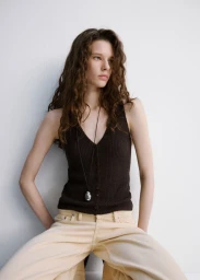 A woman wearing a dark brown ribbed tank top and pale beige trousers posing against a plain white studio background.
