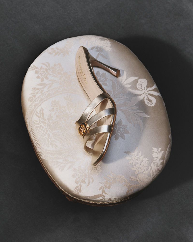 Gold high-heel sandal on a cream-colored damask cushion against a dark gray background in a studio shot, elegant and luxurious.