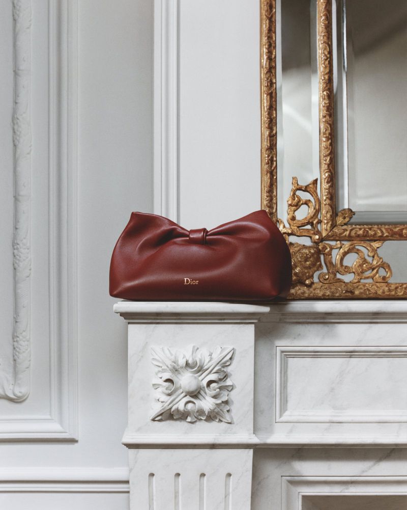 Burgundy leather Dior clutch rests on a white marble mantelpiece against a wall with elegant architectural molding and gilded mirror.
