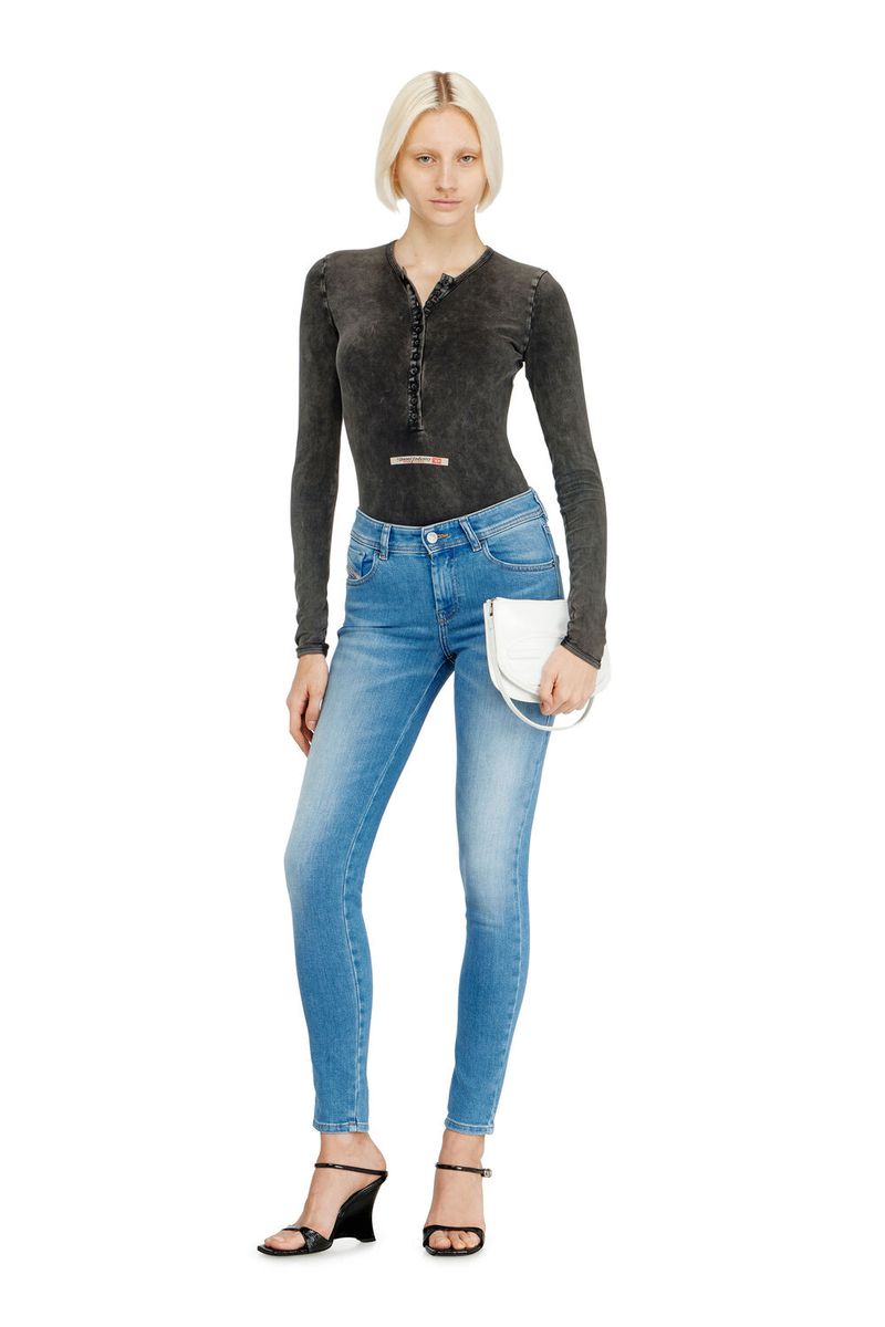 Full-length shot of a fashion model in blue jeans, gray long-sleeve top, and black sandals, holding a white purse against a white background.