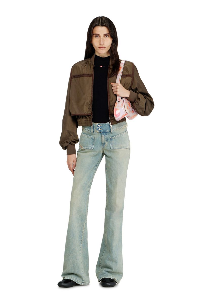 A full-length studio shot of a woman in jeans, a brown jacket, and black top, carrying a purse, set against a clean white backdrop.