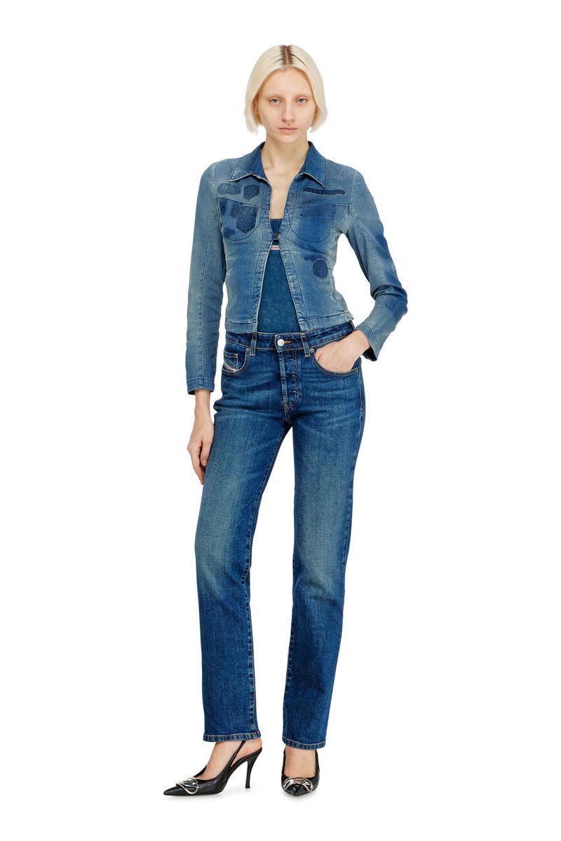 A full-length shot of a woman in dark denim jeans and a matching jacket, standing on a white background with high-heeled shoes.