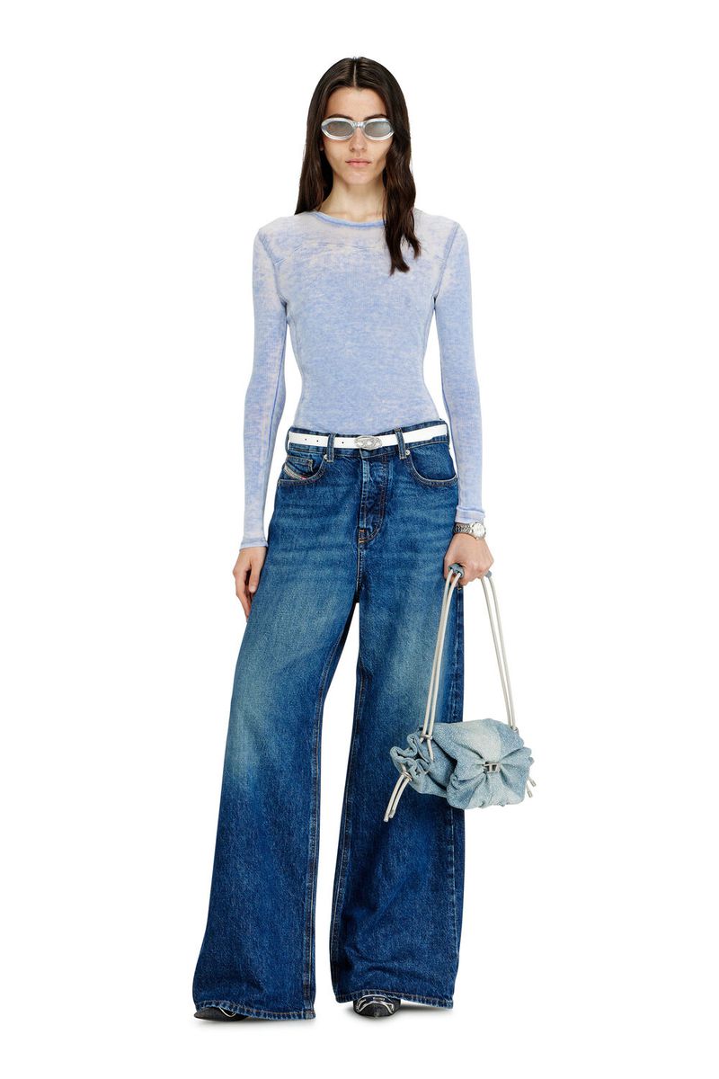 Full-length shot of a woman wearing sunglasses, long-sleeved top, wide-leg denim pants and a handbag in a white studio setting.