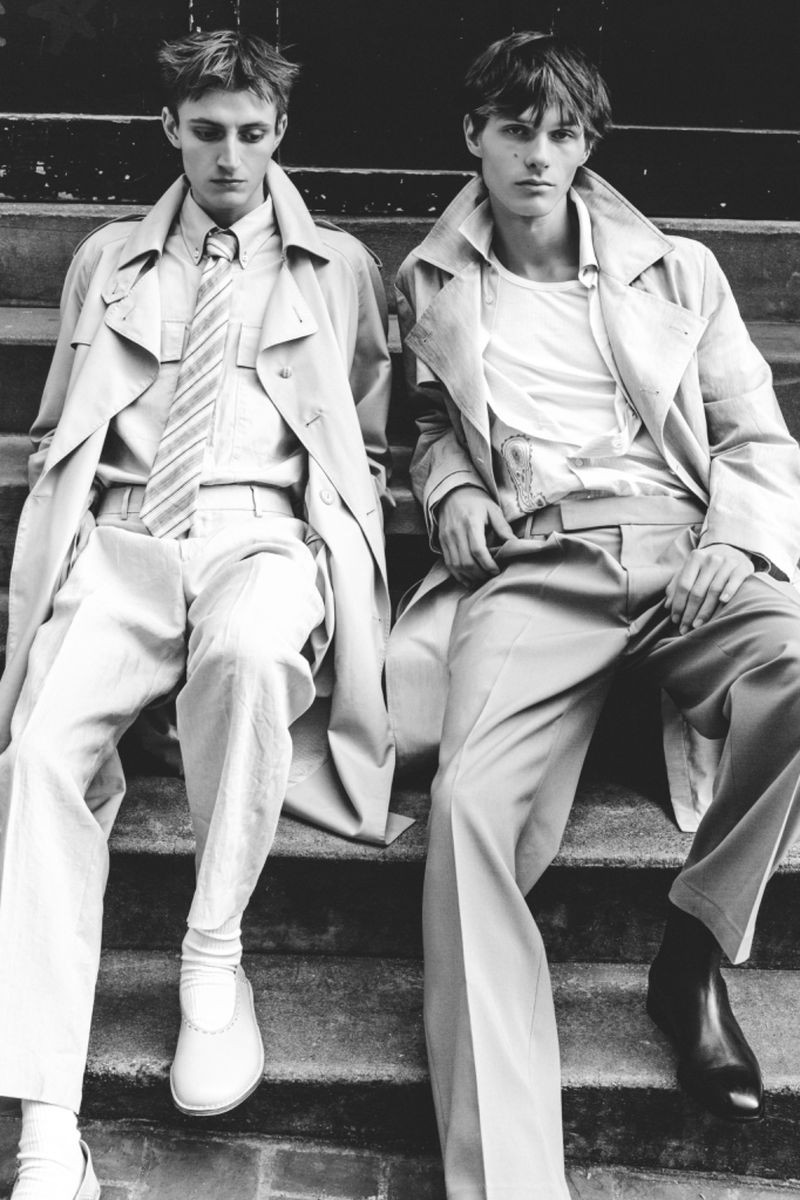 Two men in trench coats sit on steps in a black and white fashion editorial. One man wears a tie, the other wears black boots.
