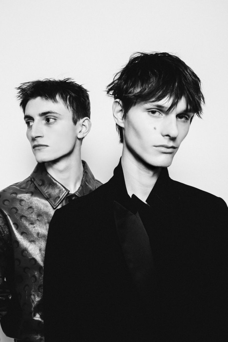 Two young adult male models in a monochromatic photo studio shot. The models are wearing contrasting jackets, in a portrait format.
