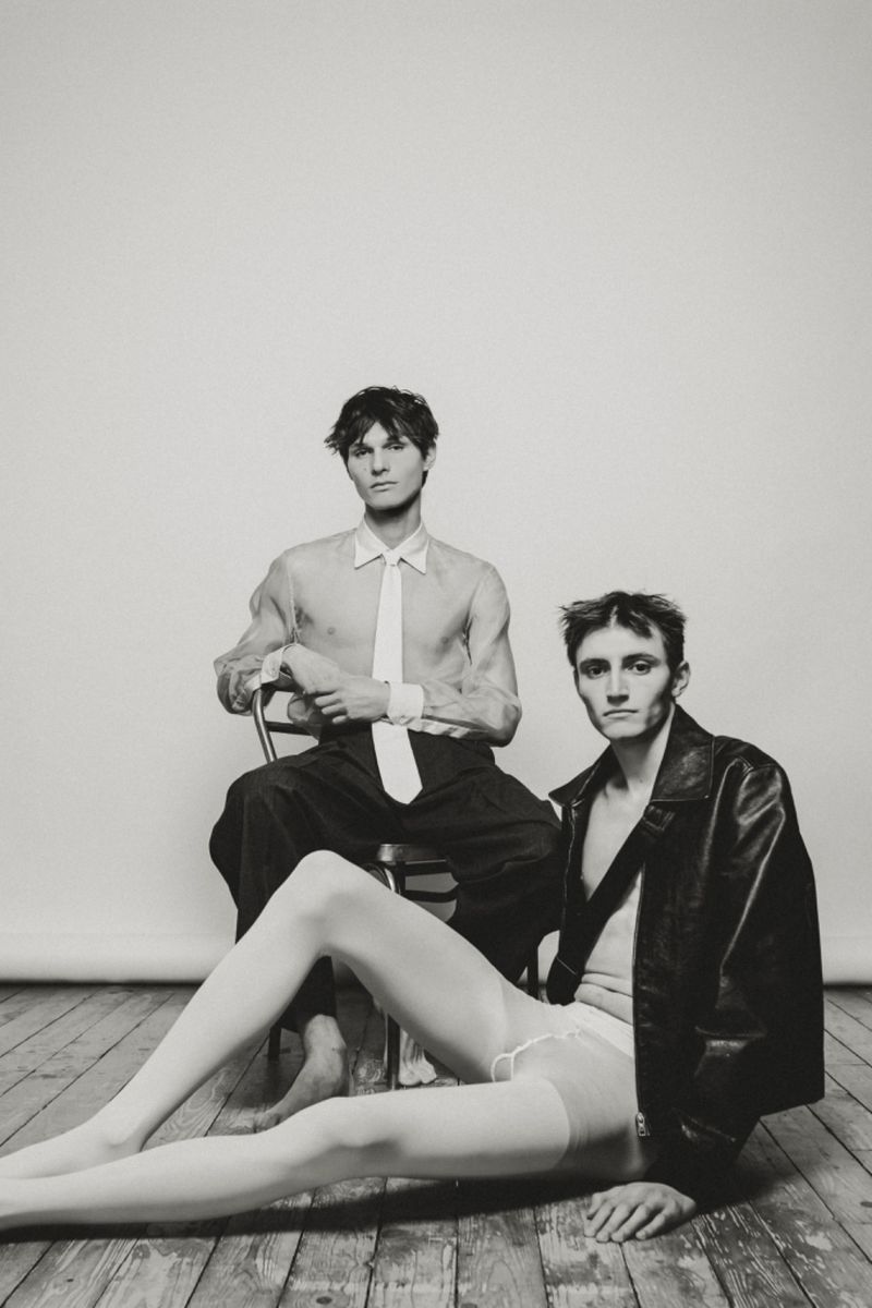 Two adult humans in a photo studio setting. One is sitting on a chair wearing a dress shirt and tie, the other reclining in hosiery and a leather jacket.