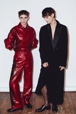 Two slender adult male fashion models in formal attire pose in front of a white wall on a wooden floor in a portrait shot.