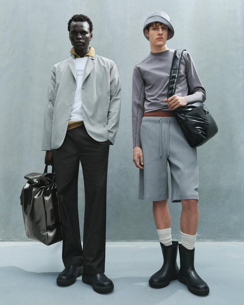 Two men in muted grey tones showcase fashion with relaxed separates, shiny black bags, and unique boots against a simple background.