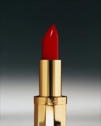 Close-up of a Celine red lipstick in a polished gold metal case engraved with Rouge Celine against a gradient studio background.