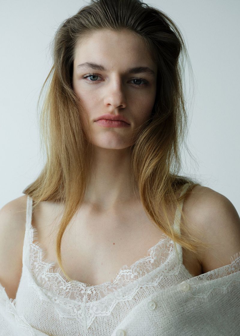 Portrait of a young adult female with long blonde hair, wearing a white lace-trimmed camisole against a neutral backdrop in natural light.
