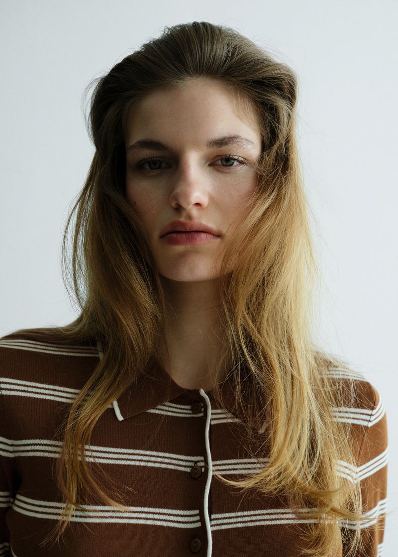 Close-up studio shot of a fair-skinned woman with long brown hair wearing a brown and white striped collared shirt looking at the camera.