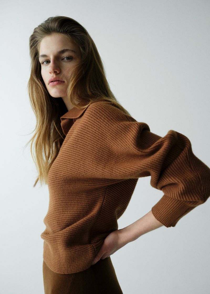 Close-up of a woman in a brown textured top with long hair, posing in front of a plain white background in a studio setting.