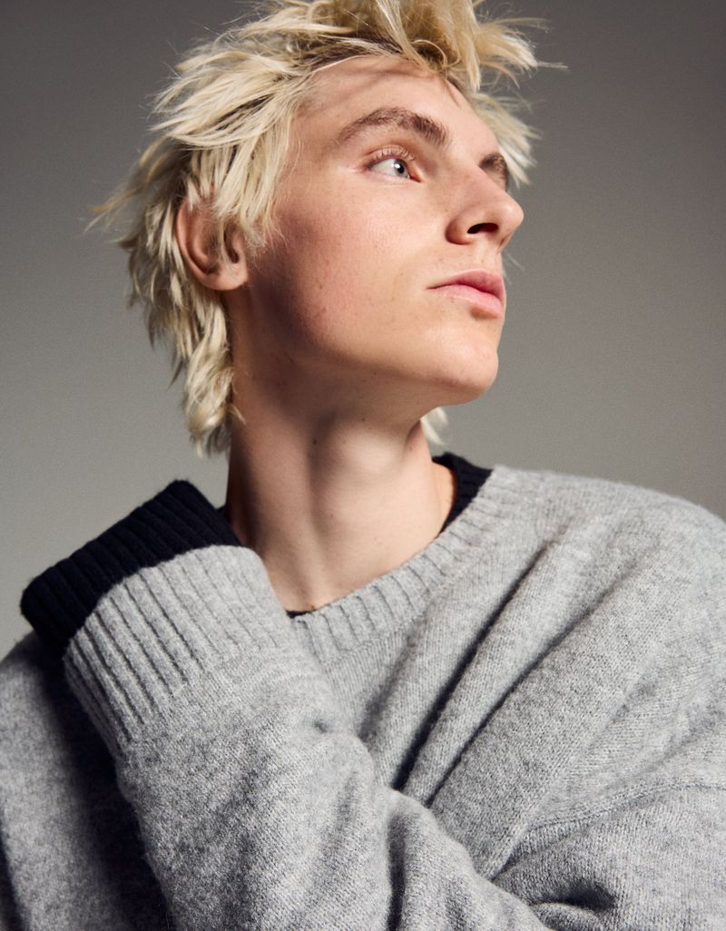 Portrait of a young adult male with blond hair in a gray sweater looking up and to the side against a neutral background.