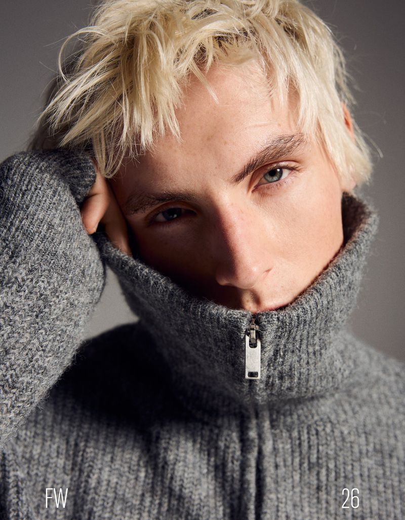 Portrait of a young adult male with light blonde hair, wearing a gray zipped cardigan, posing in front of a gray background.