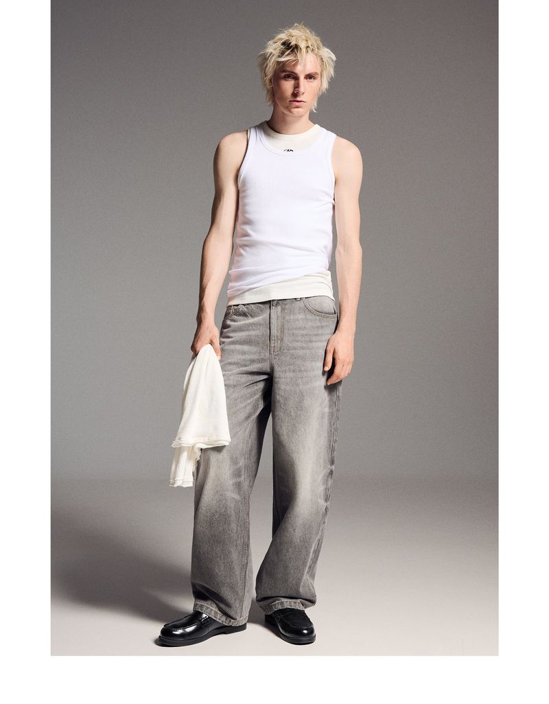 Full shot of a fair-skinned young man in a white tank top and gray jeans holding a white cloth, standing in a studio with a gray background.