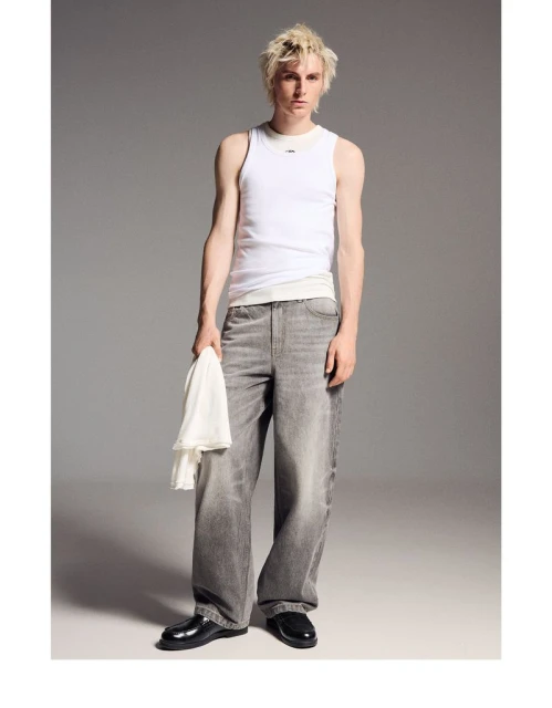 Full shot of a fair-skinned young man in a white tank top and gray jeans holding a white cloth, standing in a studio with a gray background.