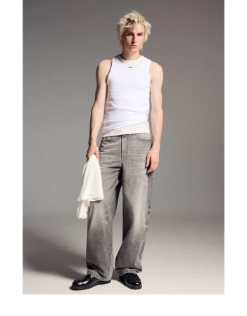 Full shot of a fair-skinned young man in a white tank top and gray jeans holding a white cloth, standing in a studio with a gray background.
