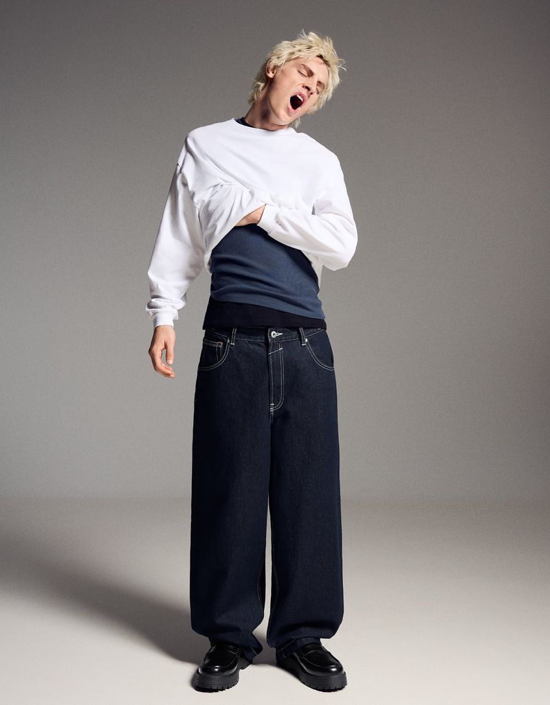 Full-length shot of a male model with blond hair, wearing a white long-sleeved shirt, dark blue pants, and black leather shoes. He's yawning.