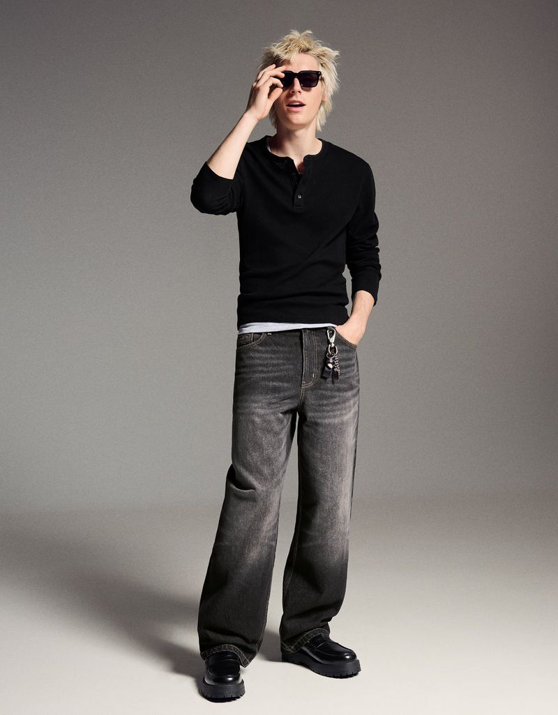 Full-length shot of a young adult man in black long sleeve shirt and grey wide-leg jeans, posing indoors in a fashion editorial style.