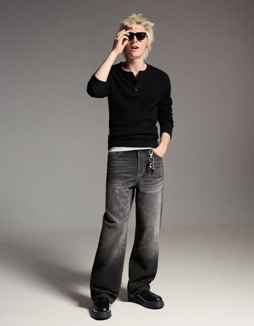 Full-length shot of a young adult man in black long sleeve shirt and grey wide-leg jeans, posing indoors in a fashion editorial style.