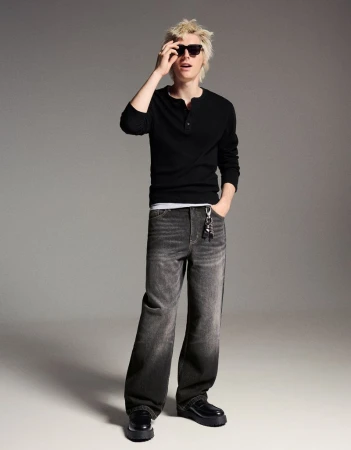 Full-length shot of a young adult man in black long sleeve shirt and grey wide-leg jeans, posing indoors in a fashion editorial style.