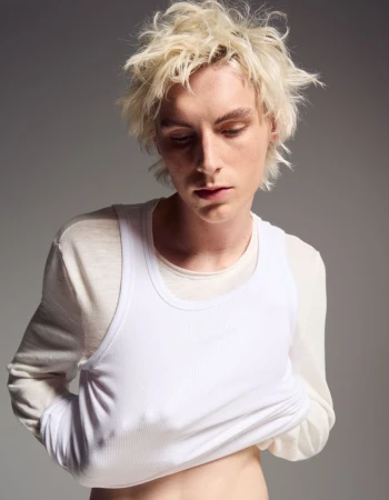 Portrait of a young adult with light blond hair wearing a white tank top and a long-sleeve shirt, set against a neutral gray background.