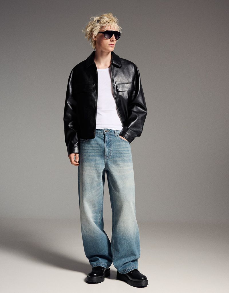 A person poses in a black leather jacket, white t-shirt, and loose, light-blue jeans in a full-length studio portrait with a gray background.