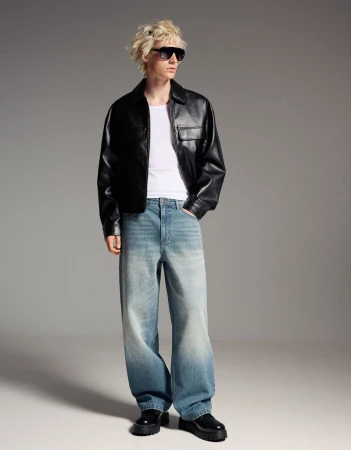 A person poses in a black leather jacket, white t-shirt, and loose, light-blue jeans in a full-length studio portrait with a gray background.