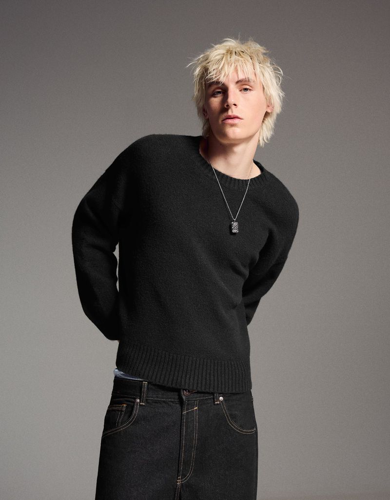 Portrait of a young adult male with bleached blond hair wearing a black wool sweater and dark denim jeans in a studio setting.