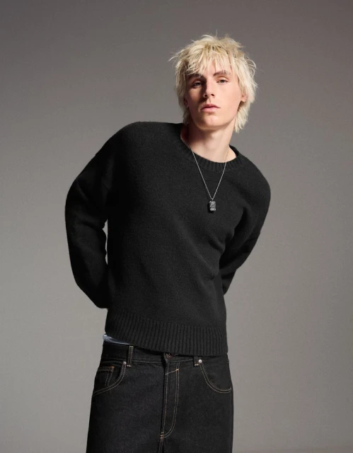 Portrait of a young adult male with bleached blond hair wearing a black wool sweater and dark denim jeans in a studio setting.