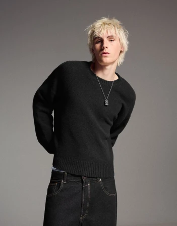 Portrait of a young adult male with bleached blond hair wearing a black wool sweater and dark denim jeans in a studio setting.