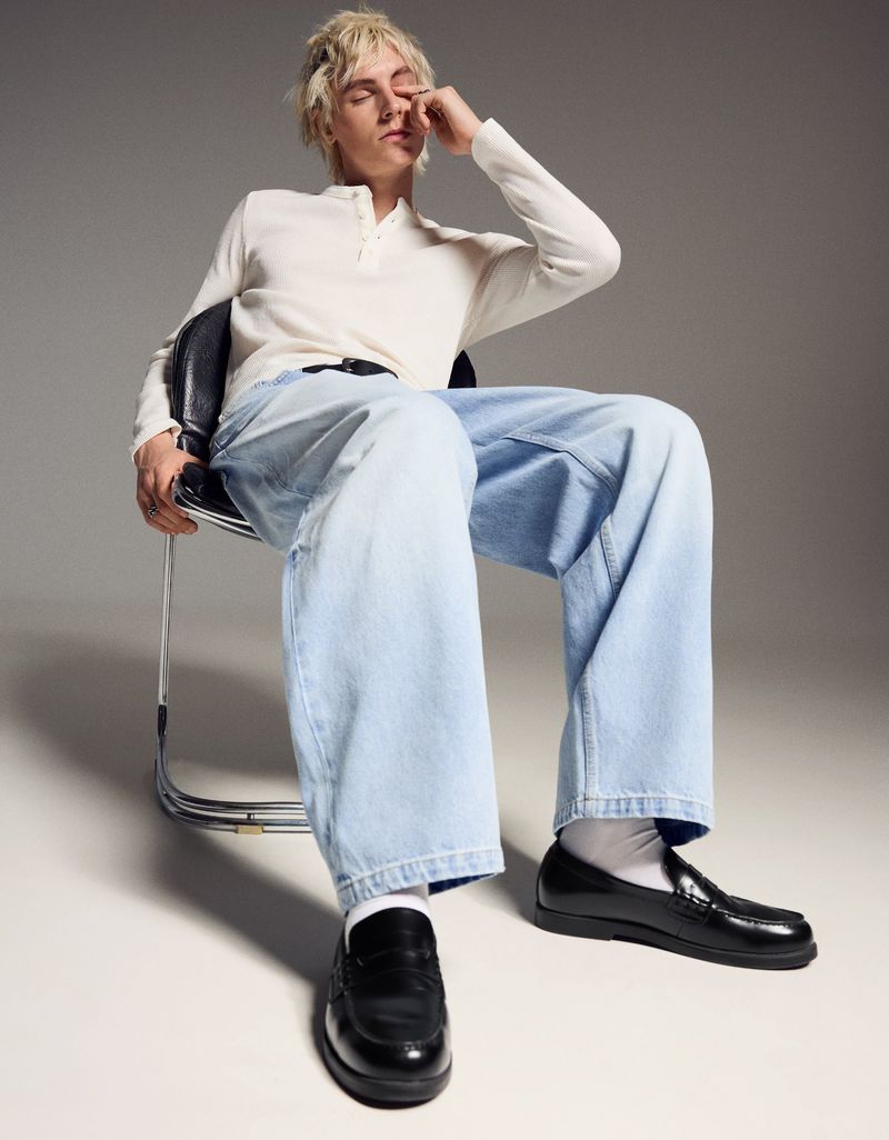 A young man with bleached blonde hair sits on a black office chair wearing a cream henley shirt and wide-leg light blue jeans with black loafers.