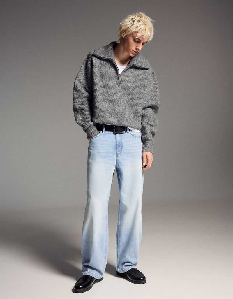 Full-length shot of a blonde male model in a grey sweater, blue jeans, and black shoes against a solid grey backdrop in a studio setting.