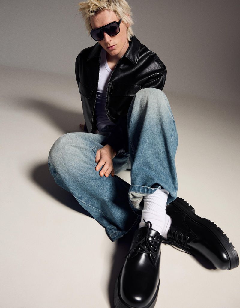 A blonde adult male model with sunglasses sits cross-legged on a light background, wearing a black jacket, white shirt, jeans, and black shoes.
