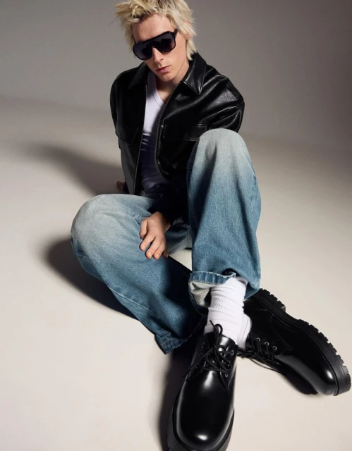 A blonde adult male model with sunglasses sits cross-legged on a light background, wearing a black jacket, white shirt, jeans, and black shoes.