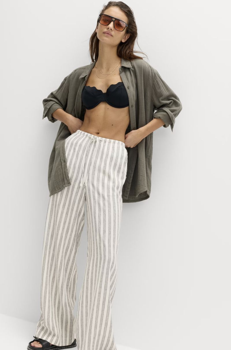 A woman in an unbuttoned green linen shirt, black bikini top, and striped linen pants poses against a white wall, showcasing casual fashion.