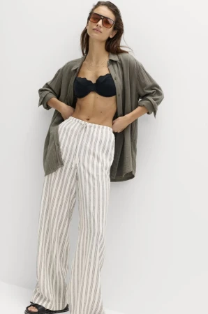 A woman in an unbuttoned green linen shirt, black bikini top, and striped linen pants poses against a white wall, showcasing casual fashion.