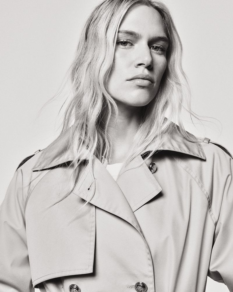 Monochrome portrait of a model with wavy blond hair wearing a light tan trench coat, looking directly at the camera with a neutral expression.