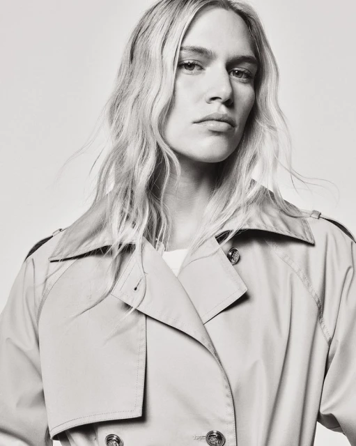 Monochrome portrait of a model with wavy blond hair wearing a light tan trench coat, looking directly at the camera with a neutral expression.