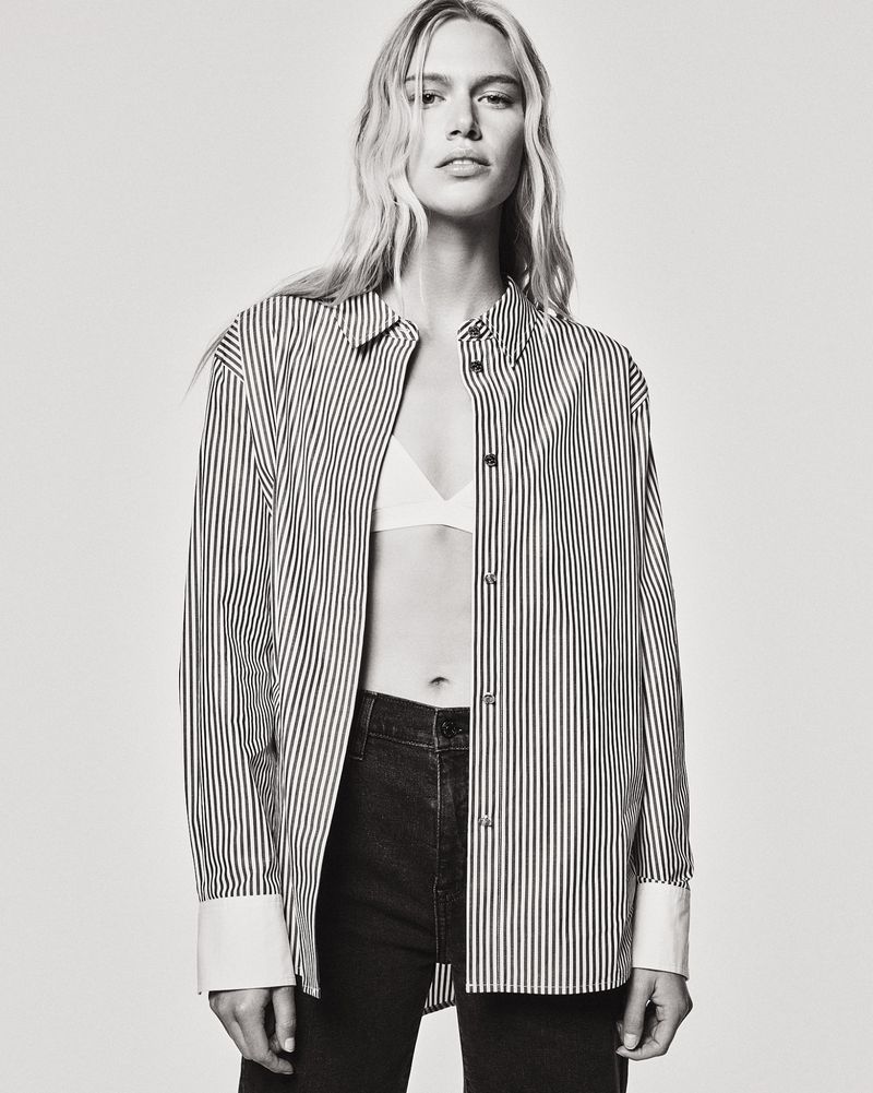 A fair-skinned woman with blonde hair poses in a photo studio wearing an oversized striped button-down shirt, jeans, and a white top.