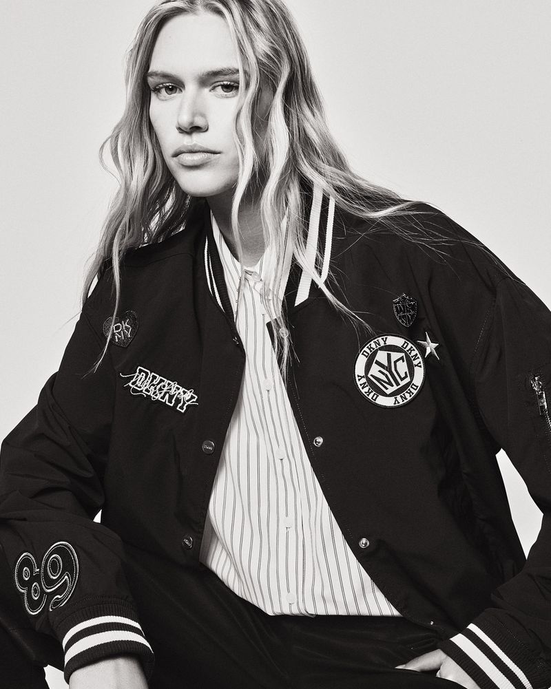 Black and white portrait of a fair-skinned person with blonde hair wearing a DKNY letterman jacket and striped button-down shirt.