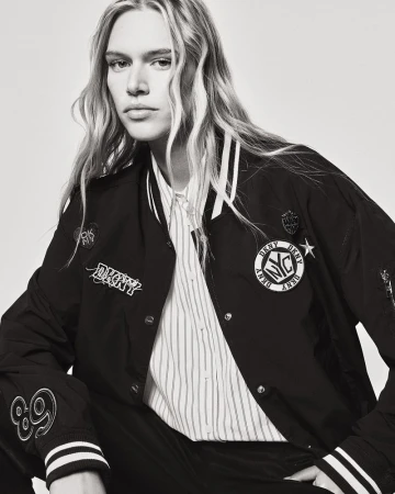 Black and white portrait of a fair-skinned person with blonde hair wearing a DKNY letterman jacket and striped button-down shirt.