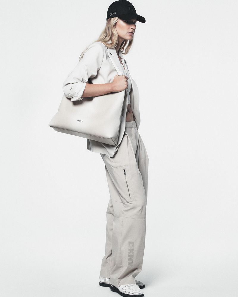 Fashion model wearing a blazer, crop top, cargo pants, sneakers, and a baseball cap holding a tote bag in a photo studio.