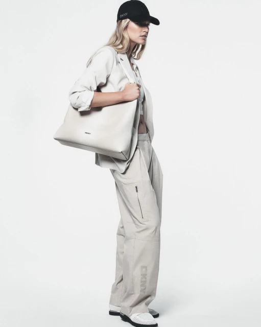 Fashion model wearing a blazer, crop top, cargo pants, sneakers, and a baseball cap holding a tote bag in a photo studio.