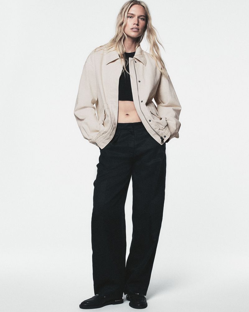 Full-length studio shot of a young adult female fashion model in a beige jacket, black crop top, and black pants against a white background.