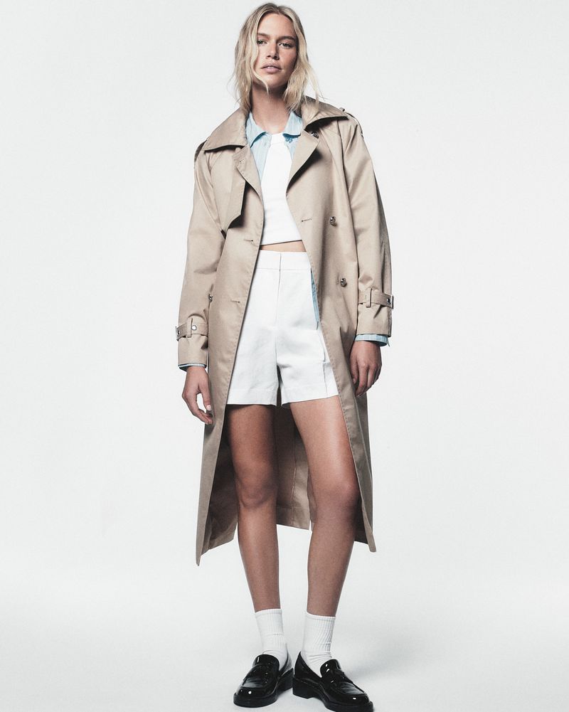 A stylish blonde woman poses in a beige trench coat, white top and shorts, with white socks and loafers against a white backdrop.